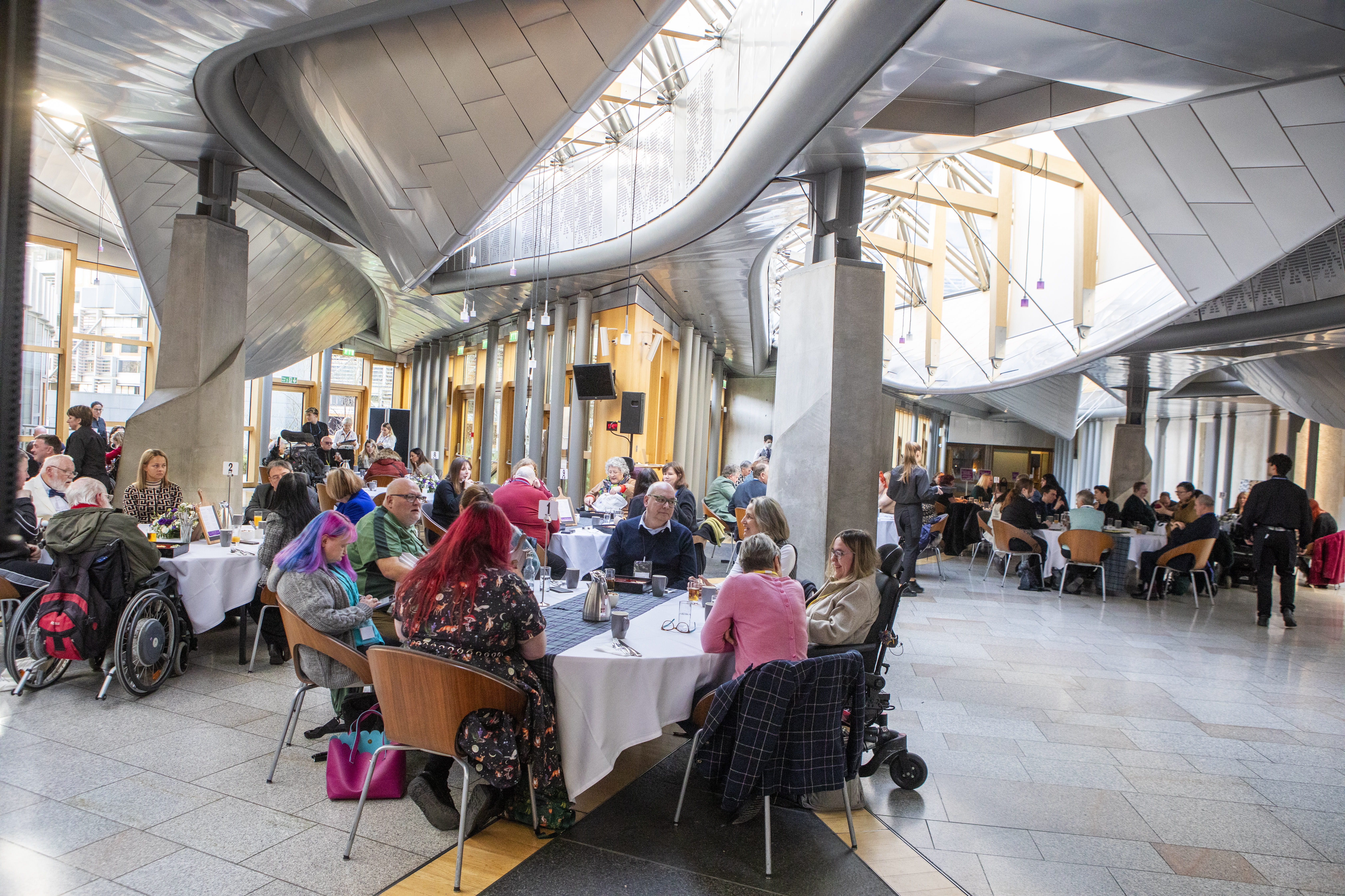 Large contemporary hall with high metal-and-glass ceiling, filled with round tables and diverse attendees, including wheelchair users, engaged in dining and conversation