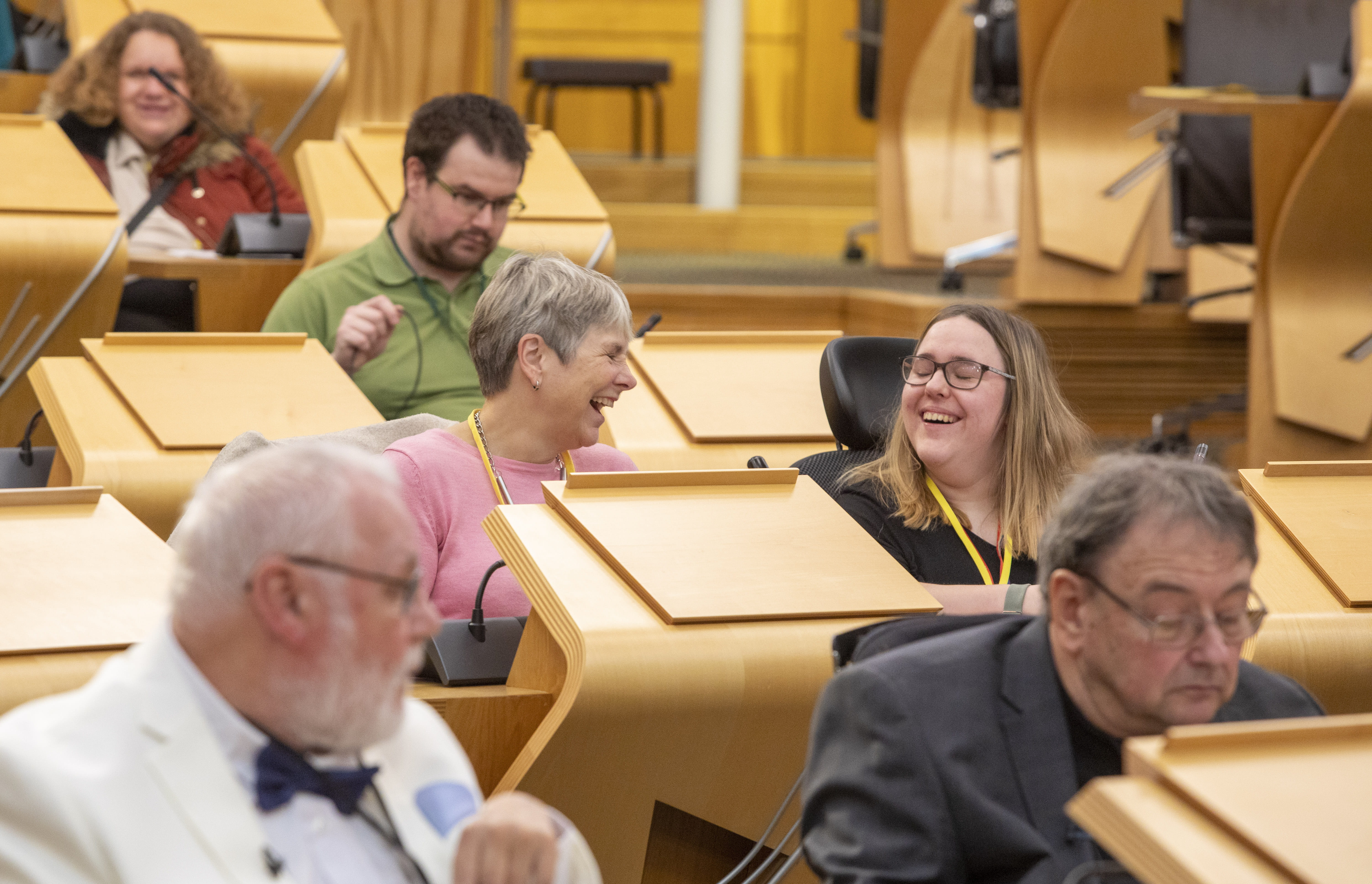 Group of people seated at wooden desks with microphones in a formal chamber, smiling and conversing during a meeting or assembly