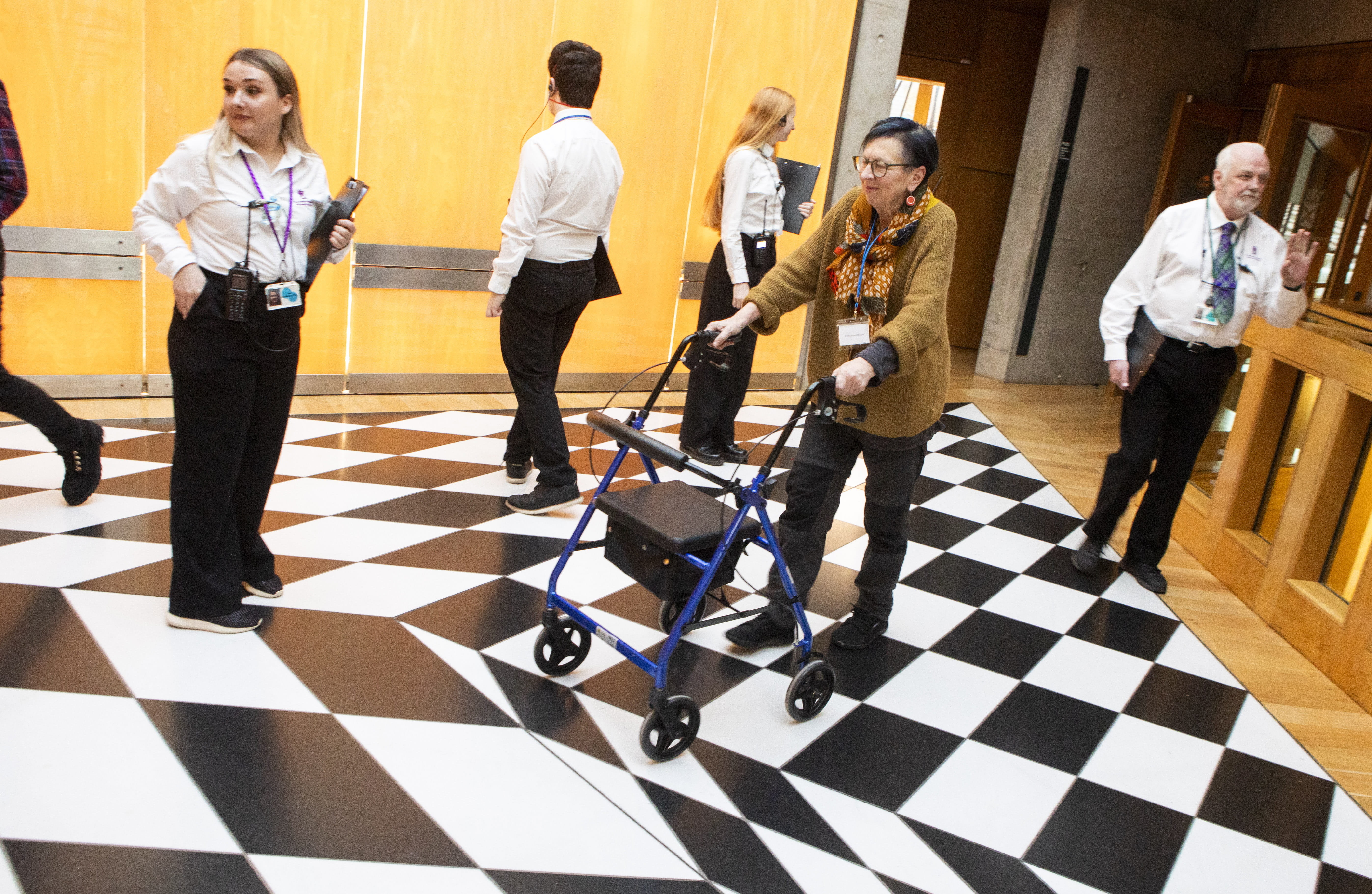 People walking across a black-and-white floor; one individual uses a blue walker in a public indoor space