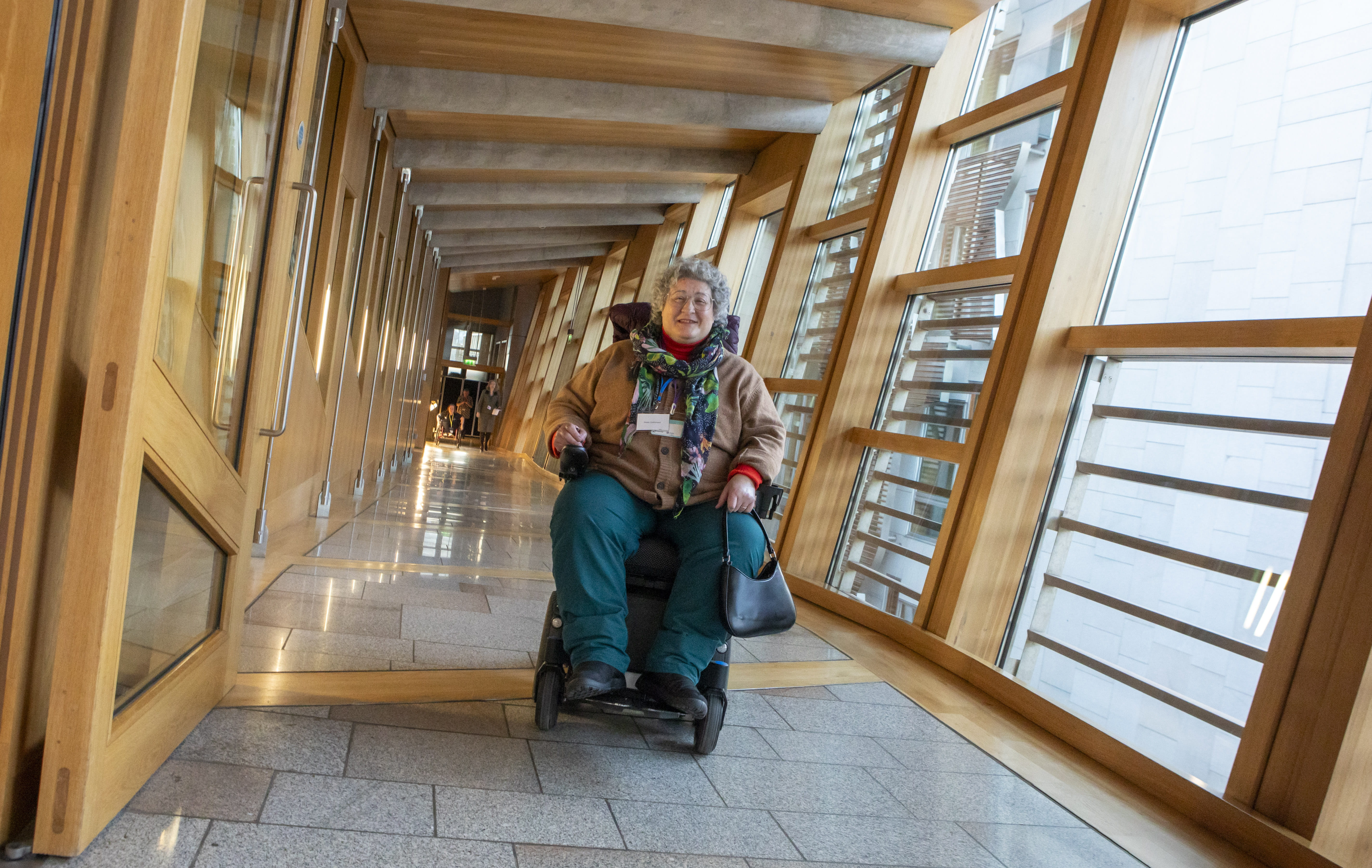 Person in motorized wheelchair moving through a sunlit hallway with wooden-framed windows and polished stone flooring