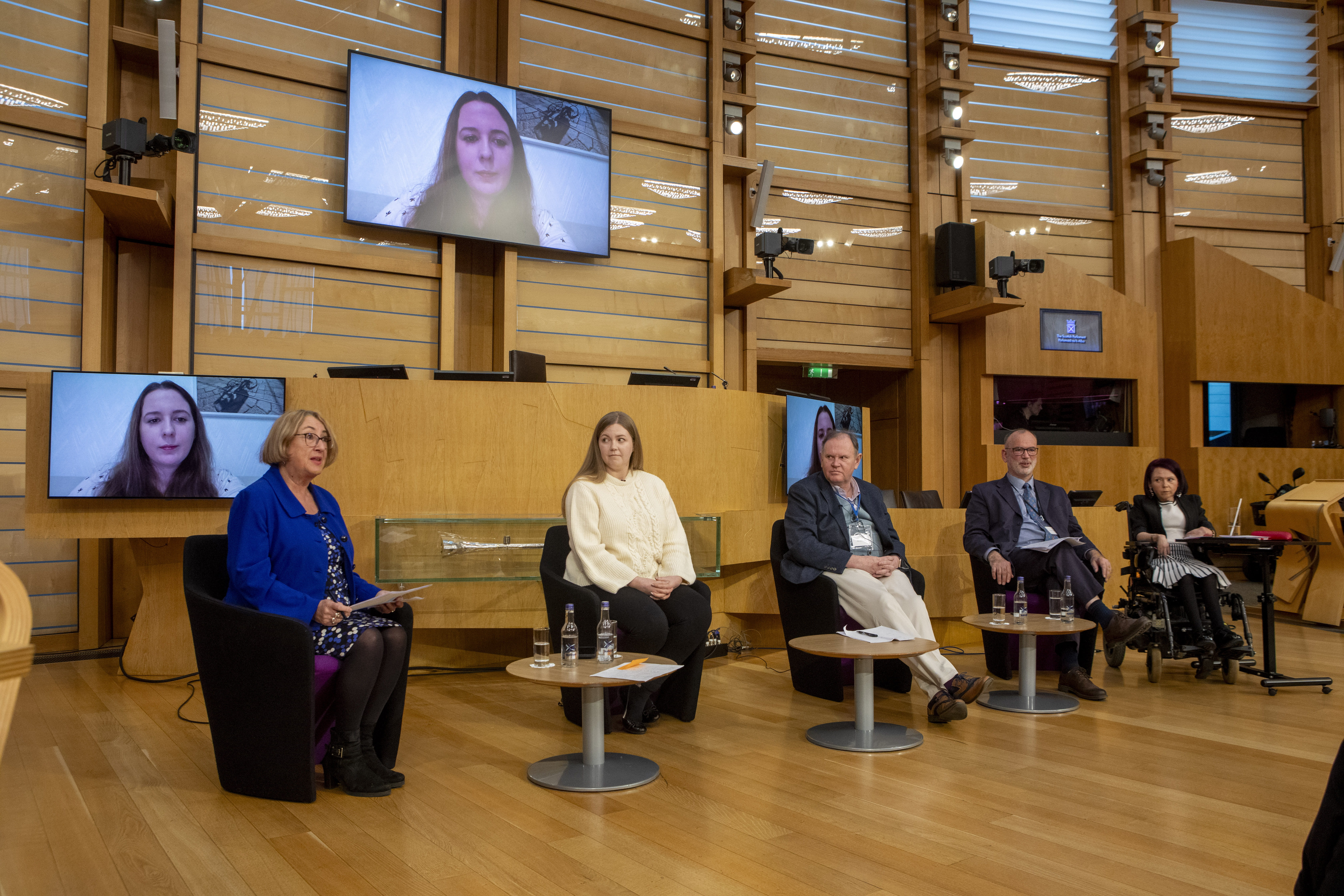 Five panelists seated on stage with water bottles and papers; hybrid event includes remote speaker on screen and visible accessibility accommodations