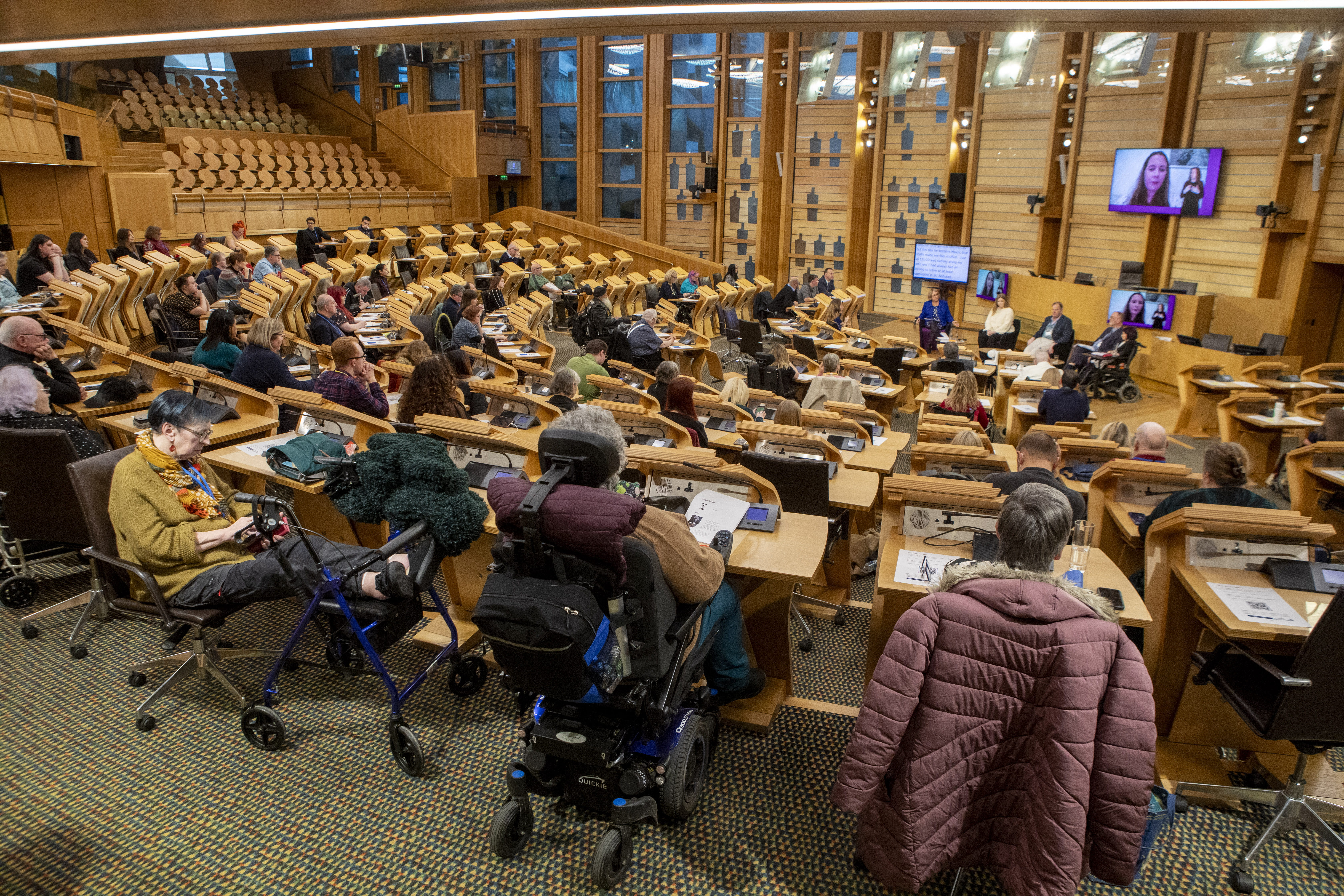Spacious chamber with semi-circular desk layout and video screens showing remote speakers; attendees include wheelchair and walker users