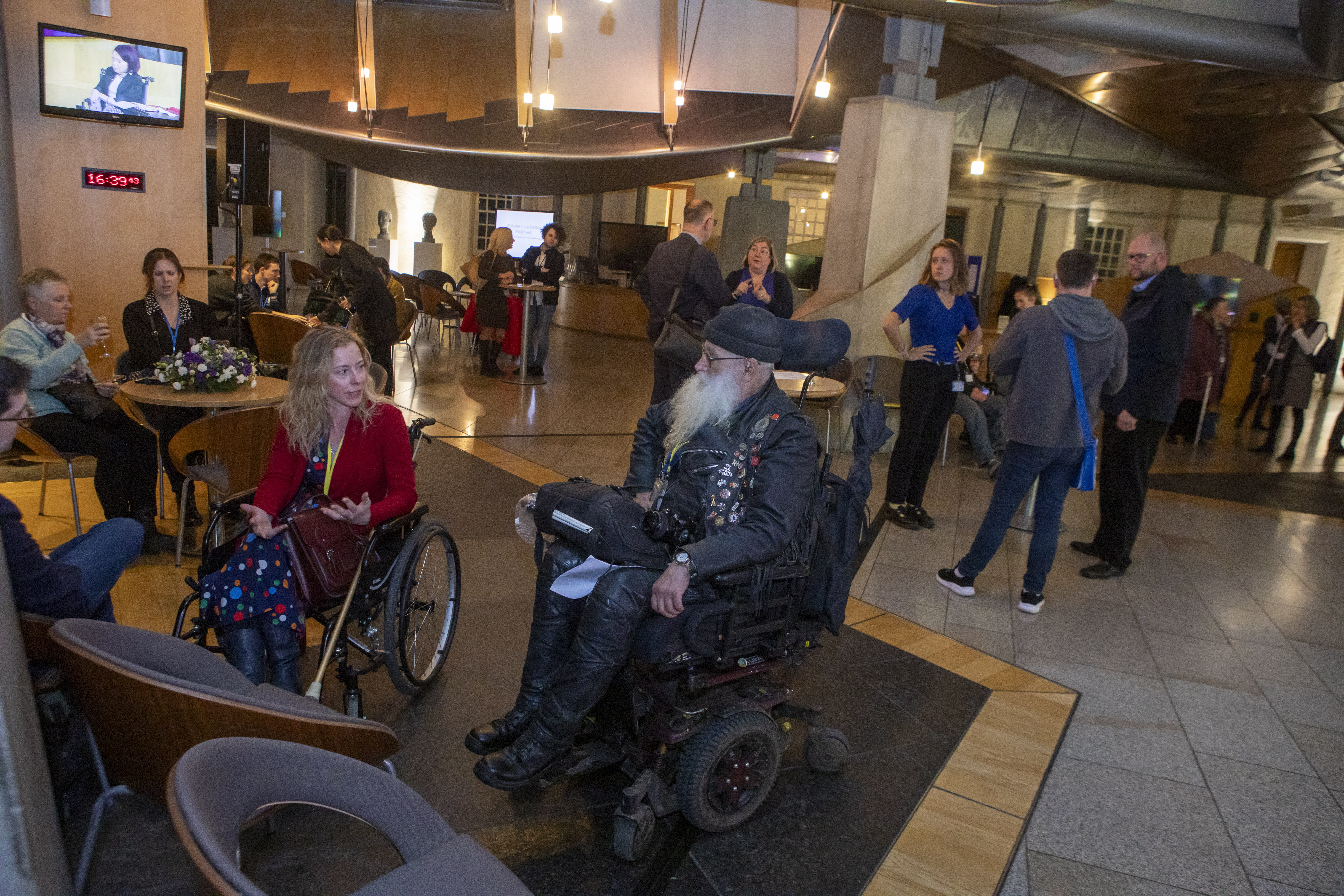 Inclusive public space with modern lighting and seating; individuals in wheelchairs actively engaged in conversation near a wall-mounted screen