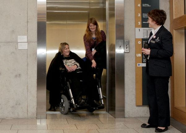 a wheelchair user and their assistant exit a lift to be greeted by a Parliament staff member
