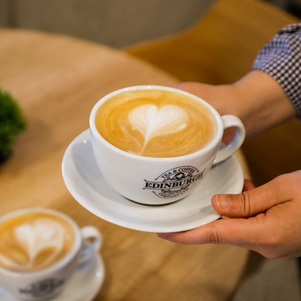 Cup of flat white coffee held above a table with another cup of coffee visible in the background.