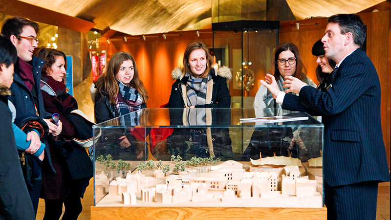 a group of visitors stands over a model of the parliament building while a tour guide talks to them