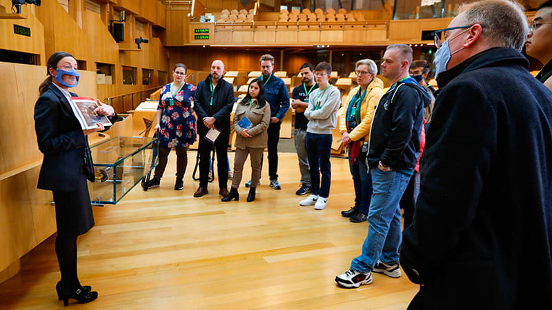A tour guide holds up a printed image to a group of visitors in the Debating Chamber